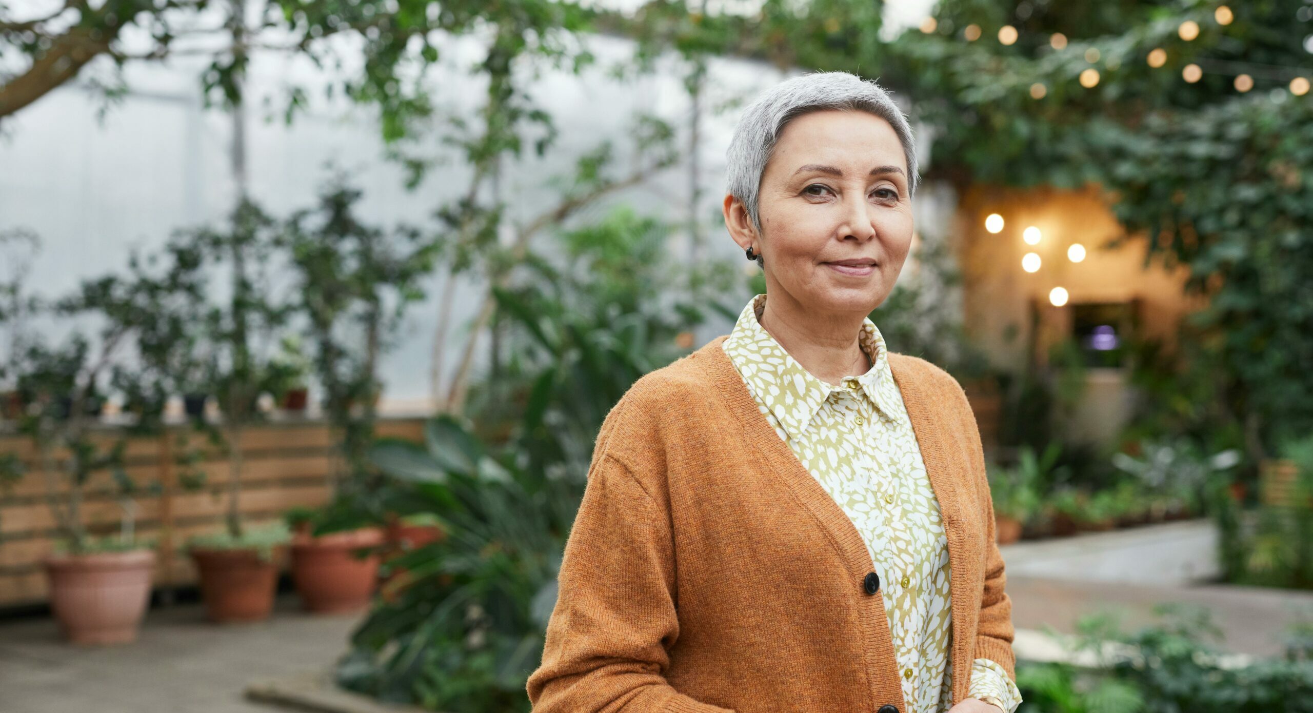 Woman with grey hair wearing a green dress and rust coloured cardigan standing in front of a greenhouse